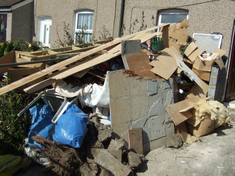 A large pile of mixed waste materials accumulated on a paved driveway, including broken pieces of wood, cardboard boxes, and concrete rubble, with some items partially covered by a white sheet and a blue plastic bag. The waste appears to be a combination of construction debris and household rubbish, situated adjacent to residential buildings visible in the background. The debris is loosely stacked, with some items leaning or resting against each other, and an assortment of cardboard packaging and damaged materials scattered around. The environment suggests an outdoor area near properties in Swiss Cottage, London, with natural daylight illuminating the scene. This image illustrates rubbish that a professional waste removal service such as Commercial Waste Swiss Cottage might collect, ensuring proper clearance and disposal of such mixed refuse from local properties.