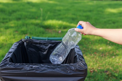 A person’s hand is shown placing a clear plastic water bottle into a black rubbish bin. The bottle is partially squeezed and features a blue cap, with visible ridges along its body. The rubbish bin is positioned outdoors on a paved area, with a lush green grassy background and diffuse natural lighting suggesting a daytime setting. The scene reflects routine waste disposal, consistent with services offered by Commercial Waste Swiss Cottage in the local area, capturing the act of discarding recyclable plastic waste as part of rubbish removal efforts. The environmental context and the practical focus on waste collection are evident, with the bin appearing to be a standard outdoor bin suitable for household or commercial use in the Swiss Cottage region, part of the NW3 postcode area.